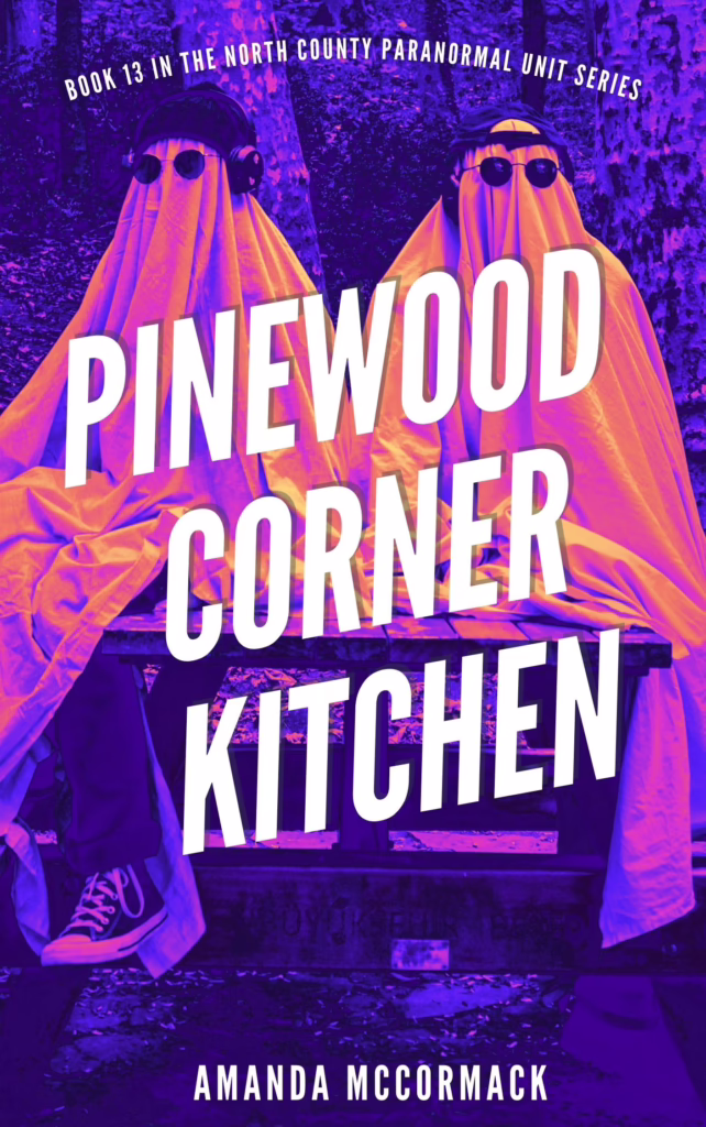 Image shows two people in white sheet ghost costumes sitting on a wooden picnic table. Both wear sunglasses and one has a beanie and headphones while the other has a backwards cap. The image is filtered in shades of pink and orange, making the ghosts bright and distinctive from the dark background of trees. Text reads Pinewood Corner Kitchen, Amanda McCormack.